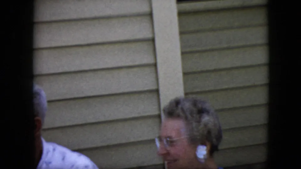 1969 California backyard home-movie frame showing an elderly woman with glasses reacting beside beige siding, with part of an elderly man at the left edge.