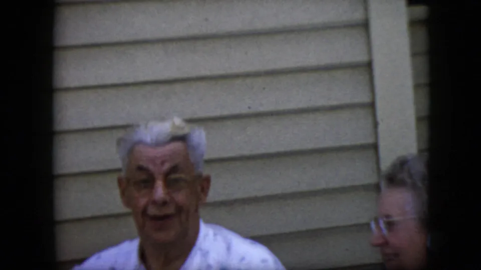 1969 California home-movie frame showing an elderly man with white hair and glasses in a light patterned shirt, with an elderly woman partly visible near beige siding.