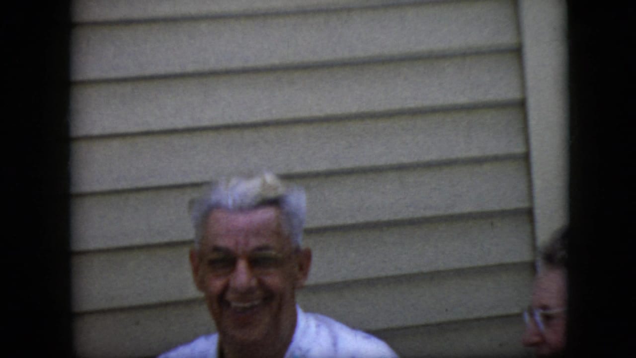 In this 1969 California home-movie frame, an older white-haired man smiles toward the camera beside beige horizontal siding while another person is partly visible at the edge.