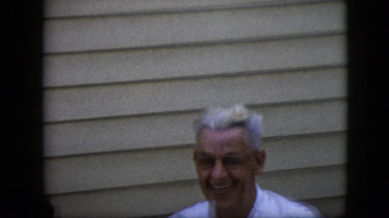 An older white-haired man in a light shirt smiles toward the camera in front of beige horizontal siding in the supplied 1969 California home-movie footage.