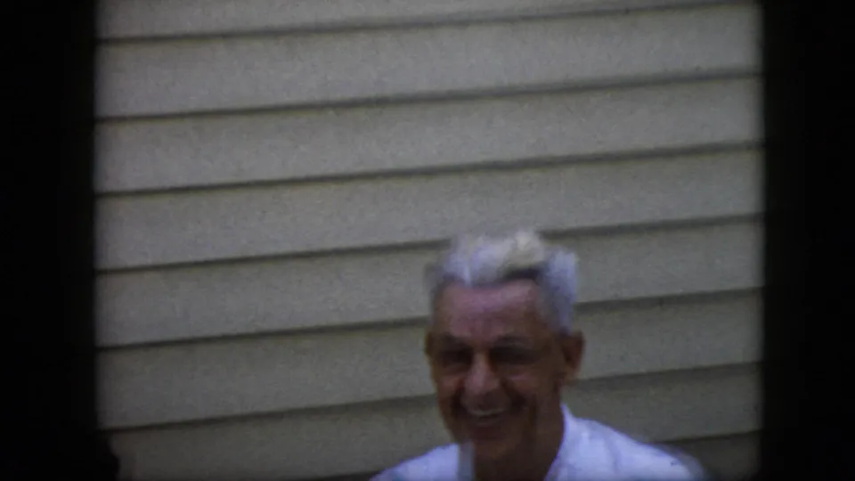 An older white-haired man in a light shirt smiles toward the camera in front of beige horizontal siding in the supplied 1969 California home-movie footage.