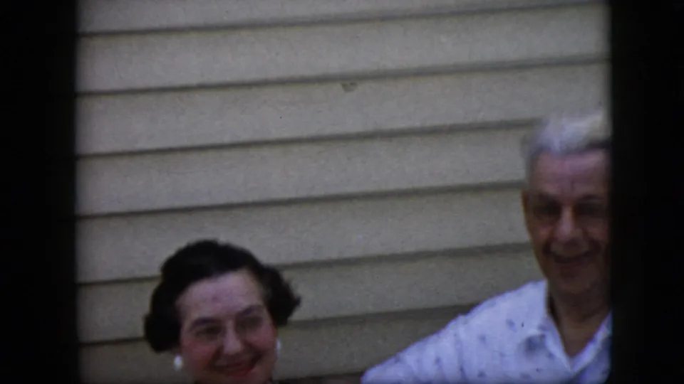 A dark-haired woman wearing earrings and an older white-haired man smile near beige horizontal siding in this late-1960s California home-movie frame.