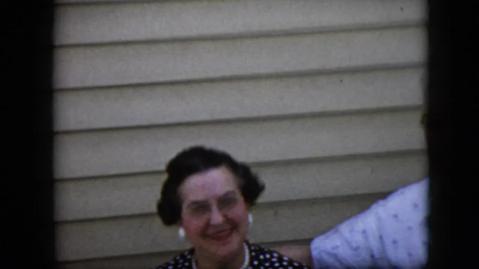 A dark-haired woman wearing earrings and a dark patterned top smiles toward the camera, with part of an older man's light shirt visible at the right edge.