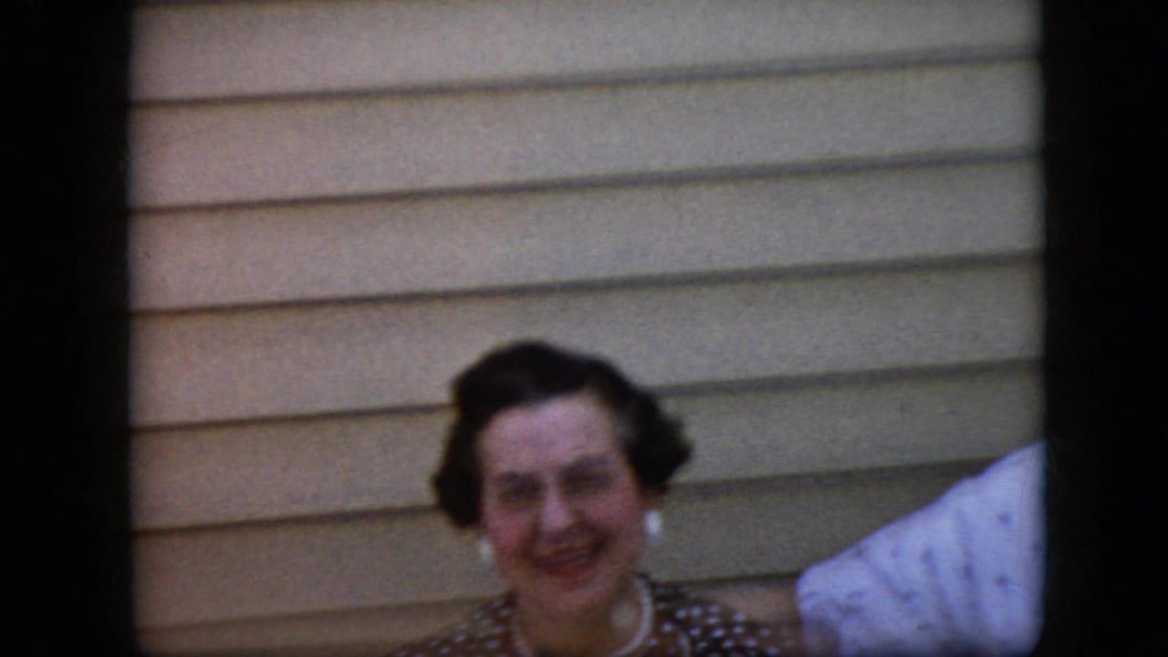 In supported 1969 California home-movie footage, an adult woman wearing a dark polka-dot dress and pearl-style jewelry smiles toward the camera beside beige horizontal siding.
