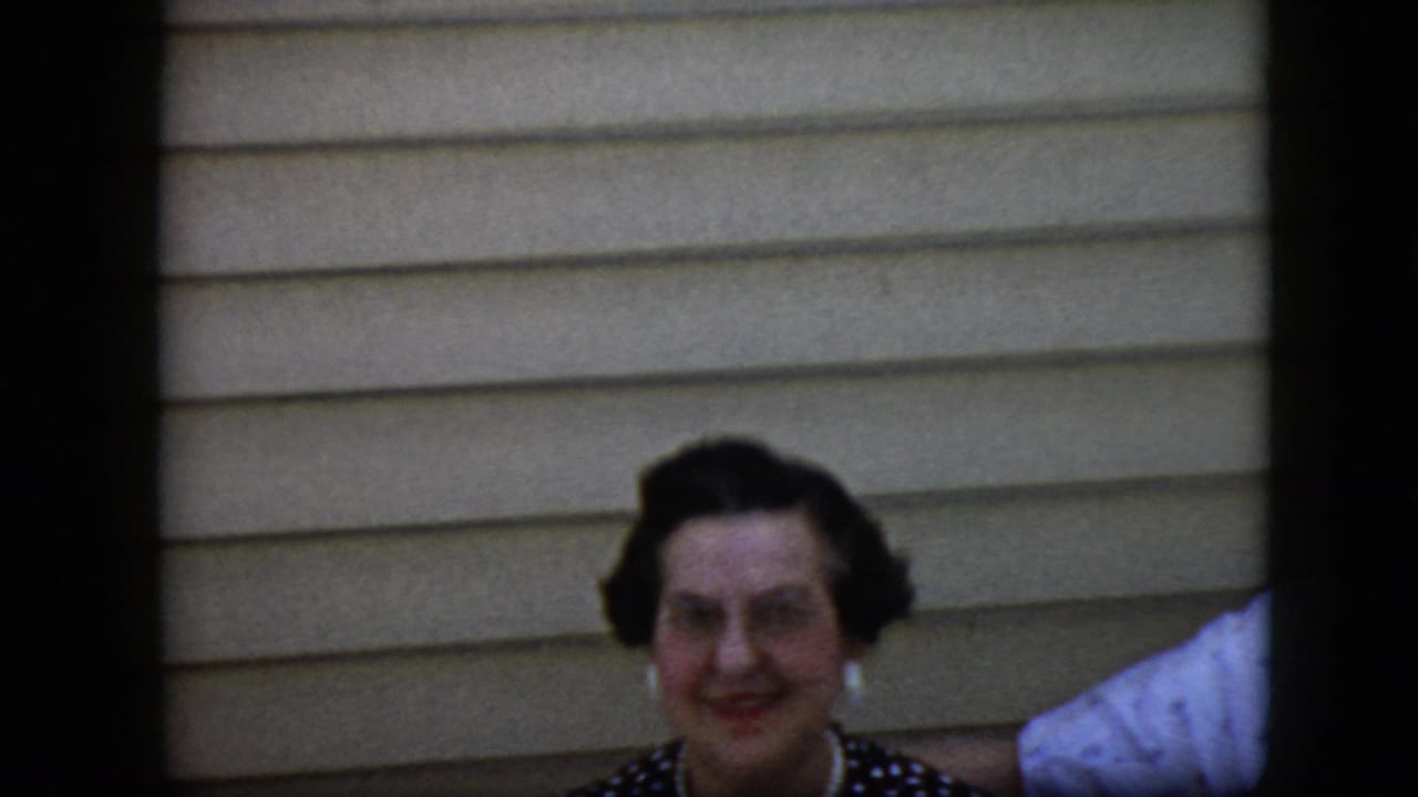 Woman smiles at the camera with beige siding behind her and partial white clothing at the right edge.