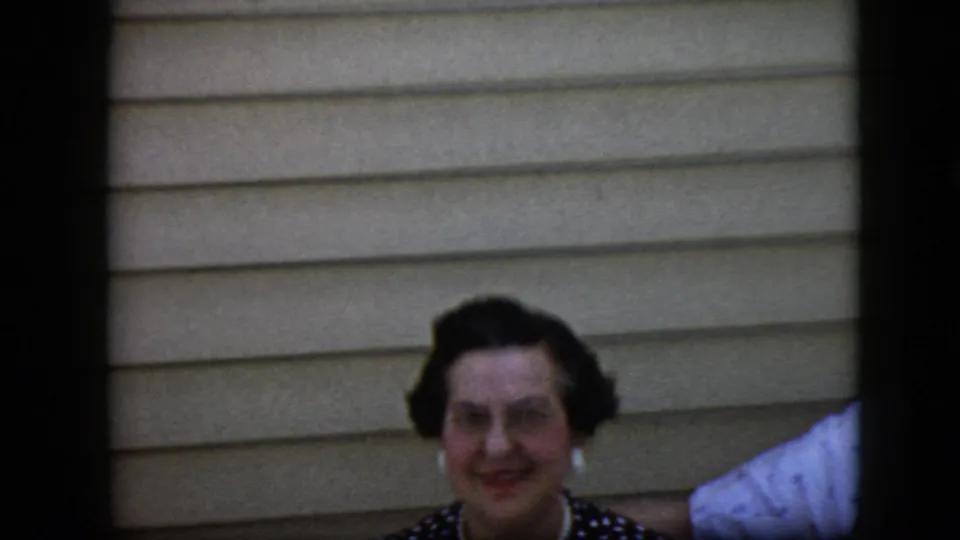 The woman remains centered in the 1969 California home-movie frame, smiling in front of beige horizontal siding with a partial white garment visible at the right edge.