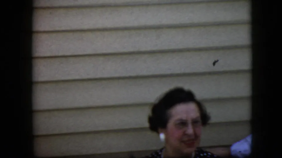 The woman shifts lower and slightly sideways in the soft 8mm frame, with beige horizontal siding filling most of the background.