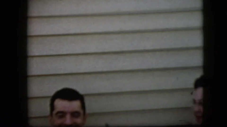 An adult man briefly appears low in the 1969 California home-movie frame, with beige siding behind him and the woman only partly visible at the right edge.