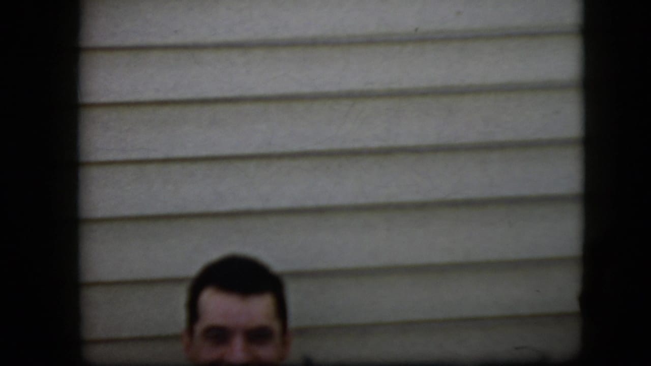 Smiling man's head partly visible against horizontal house siding, framed by dark edges.