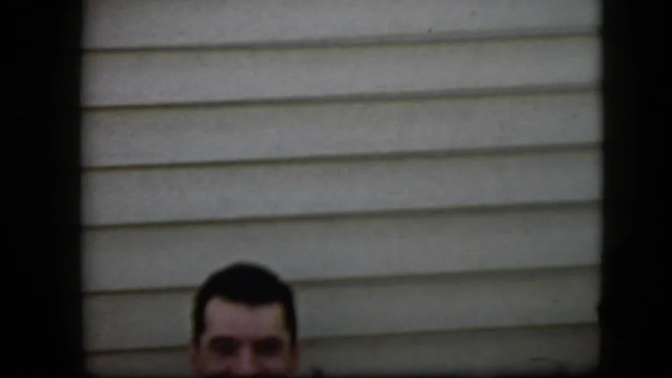 In this 1969 California home-movie frame, the upper part of a smiling young man's face appears near light horizontal house siding with dark border areas around the frame.