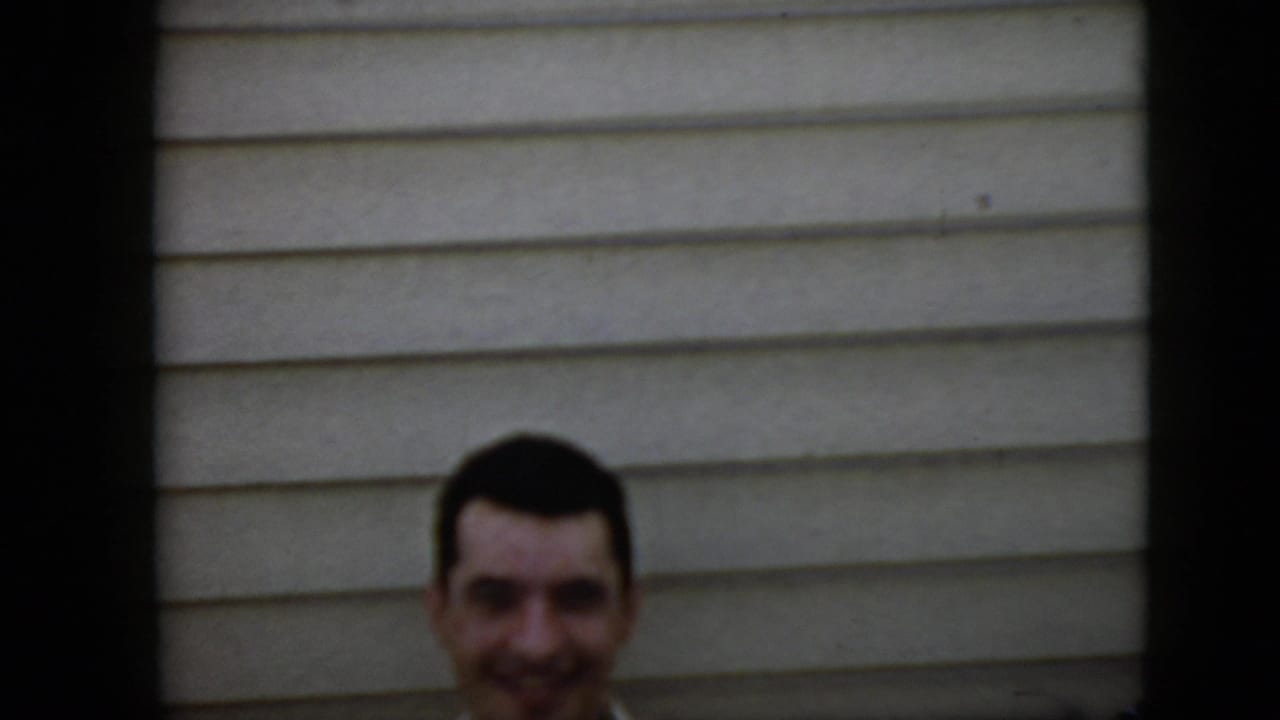Smiling man seen from the neck up beside light horizontal siding.