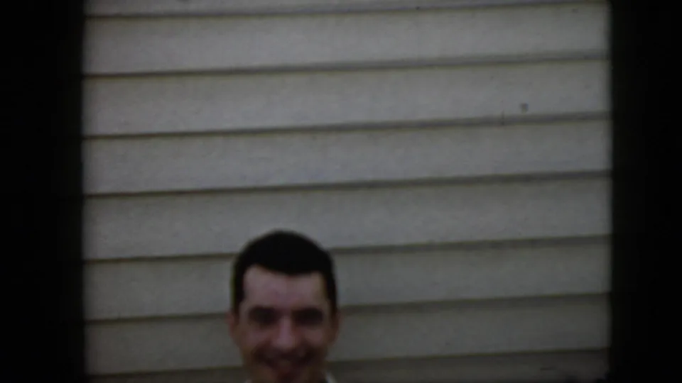 In this 1969 California home-movie frame, a smiling young man with dark hair and a mustache faces the camera near light horizontal house siding, with a dark vertical divider in the foreground.