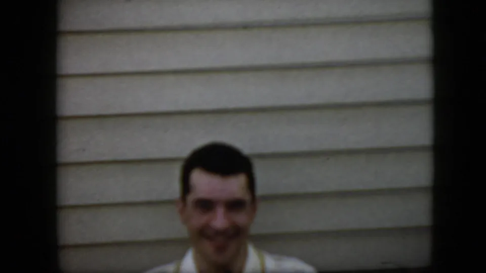 In this 1969 California home-movie frame, the man is shown from the shoulders up in a light collared shirt, smiling toward the camera against light-colored horizontal siding.