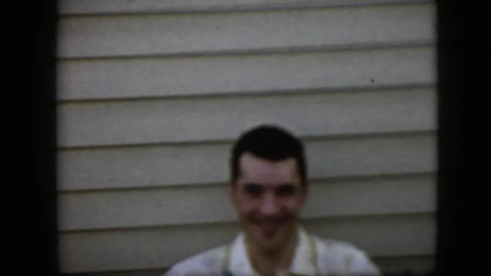 In this 1969 California home-movie frame, a smiling young man with dark hair and a light collared shirt faces the camera beside light horizontal house siding, with dark borders and soft film focus.