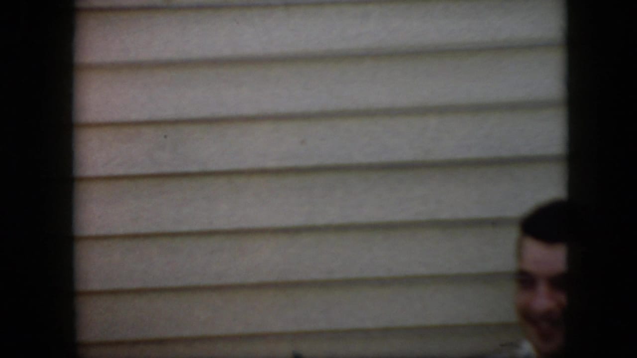 Partial view of a younger dark-haired man smiling near house siding.