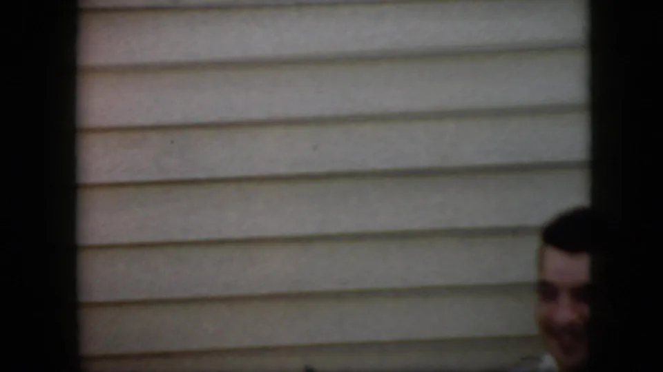 A younger adult man with dark hair appears low in the frame outside a light-sided house in this 1969 California home-movie frame.