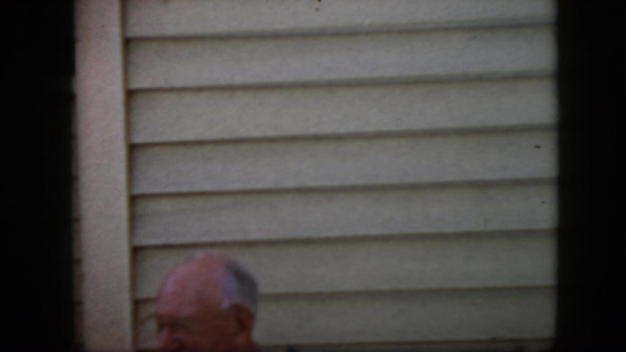 Older bald man with glasses partly visible in front of house siding.