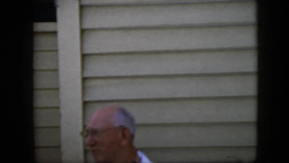 The older man with glasses appears in profile beside pale vertical trim and light horizontal siding in a candid 8mm home-movie frame.