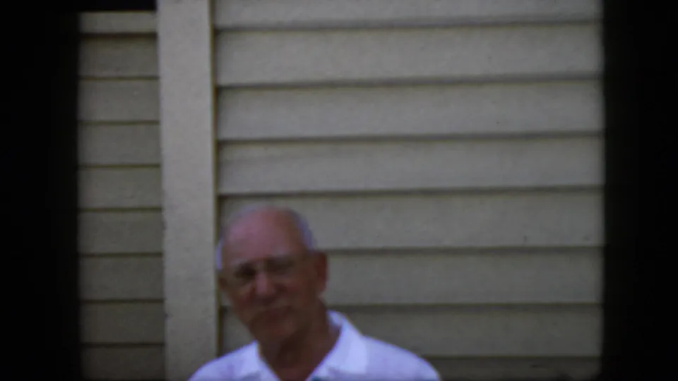 An older bald man in glasses and a light collared shirt looks toward the camera outside a light-sided California house, matching the 1969 clip metadata.