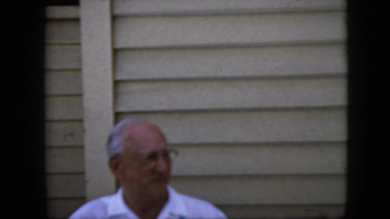 Elderly man wearing glasses and a white collared shirt appears beside beige horizontal siding in 1969 California home-movie footage, framed by dark vertical borders.