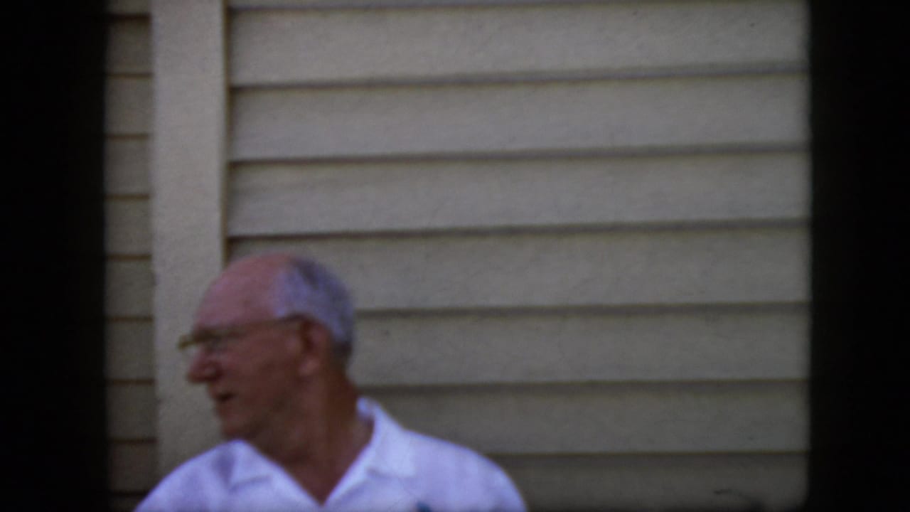 Elderly man with glasses turns in profile near beige house siding.