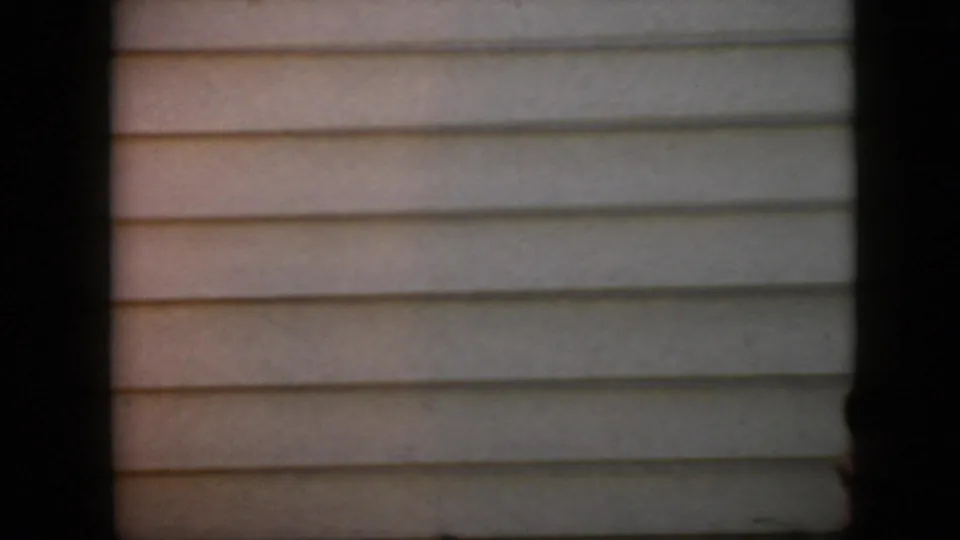 Mostly empty view of beige horizontal siding and a low ledge, framed by dark vertical borders in the same 1969 California home-movie shot.