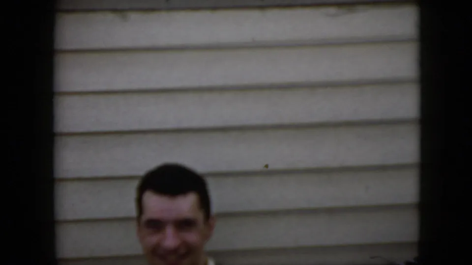 A smiling adult man appears partly framed above a low edge against beige horizontal siding in the 1969 California home-movie footage.