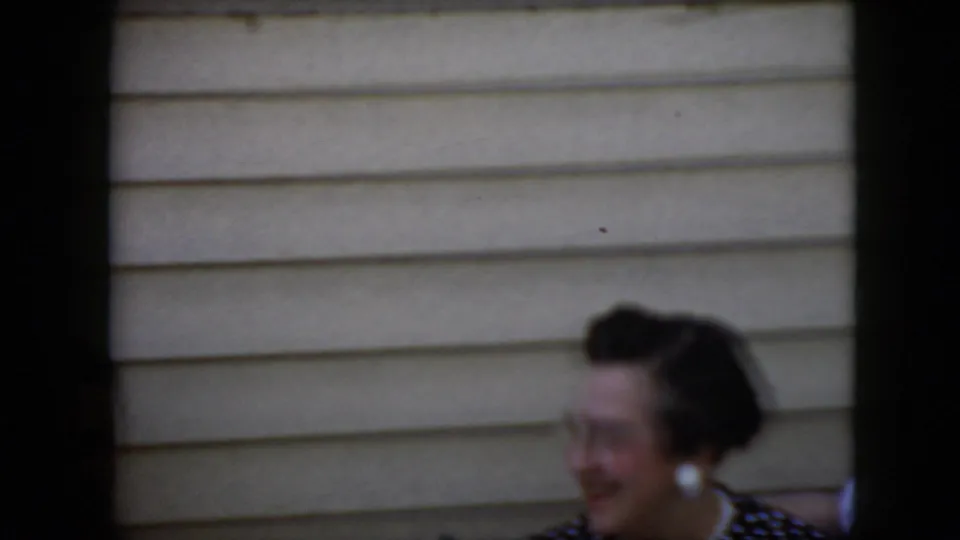 A woman with dark hair, white earrings, red lipstick, and a dark polka-dot dress smiles near beige siding in supported 1969 California home-movie footage.