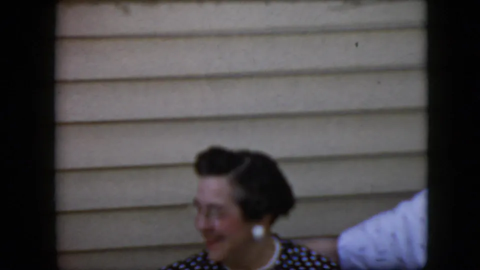 A smiling woman in a dark polka-dot dress and white earrings is seen beside a partial white sleeve against beige siding in the 1969 California home-movie frame.