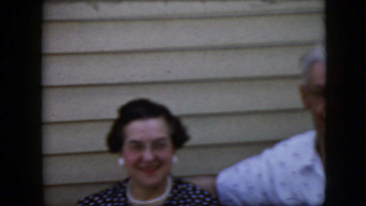Smiling woman in a dark polka-dot dress beside a partially visible man against beige siding.