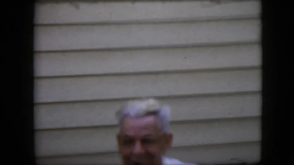 An older white-haired man is partly visible near the lower edge of the frame, with beige exterior siding behind him in the 1969 California home-movie footage.