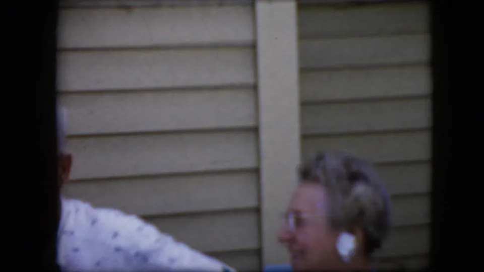 An older woman wearing glasses and light earrings appears in profile near a beige-sided wall, with another adult's white patterned shirt cropped at the edge.
