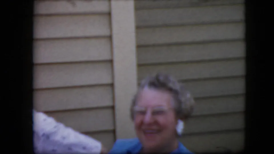 An older woman in blue clothing smiles toward the camera near a beige-sided exterior wall, with part of another adult cropped at the left edge.