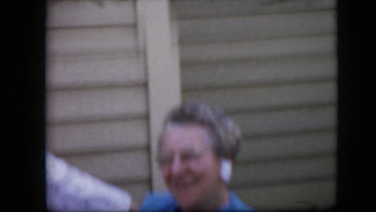In 1969 California home-movie footage, an elderly woman with glasses smiles near light horizontal siding while another figure is partly visible at the edge.