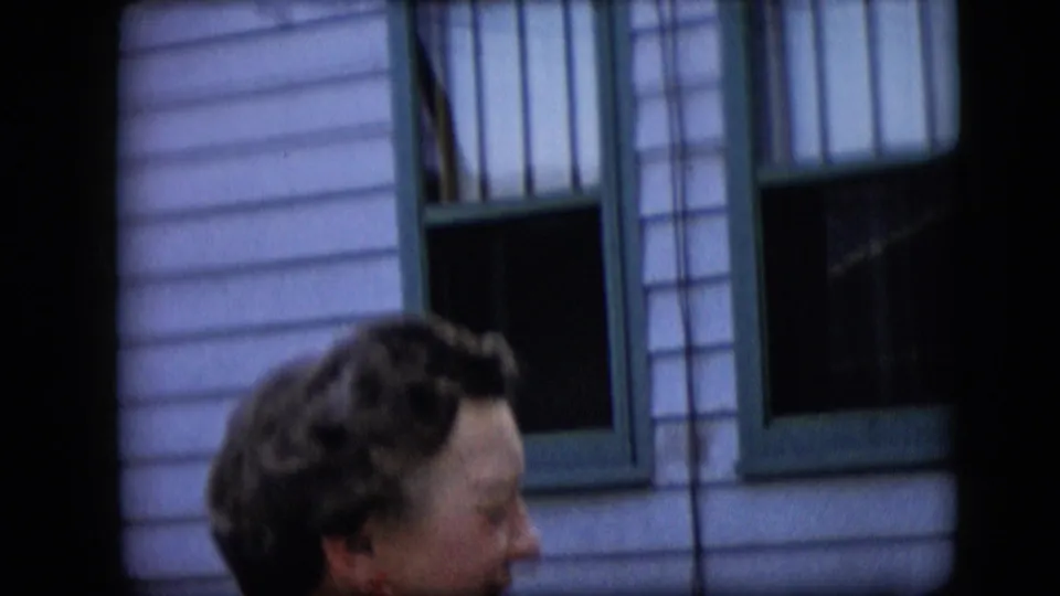 The frame shows an elderly woman in profile at the lower edge of the frame beneath green-trimmed windows and light horizontal siding.
