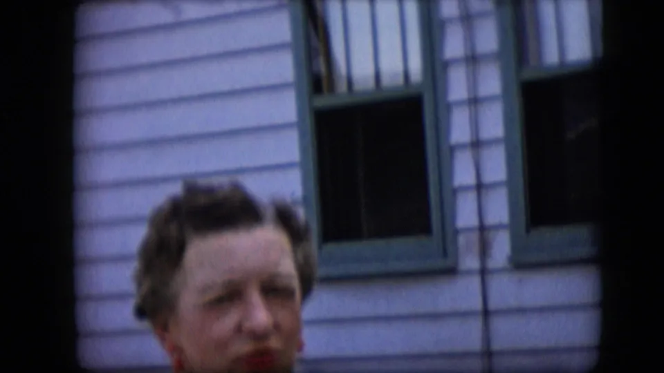 An elderly woman with dark wavy hair faces toward the camera in soft focus, framed below green-trimmed windows on a light-sided house exterior.
