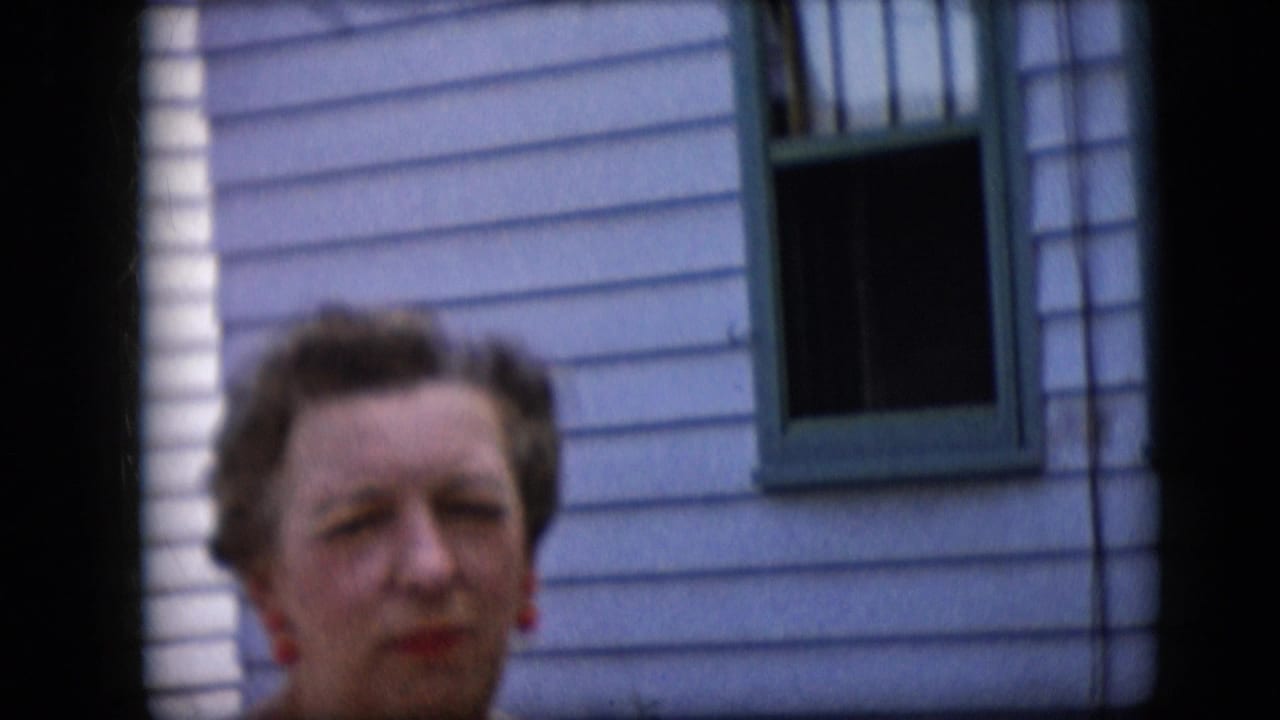 Mature woman with styled hair and red earrings appears outdoors in 1969 California home-movie footage, standing before light horizontal siding and a green-framed window.