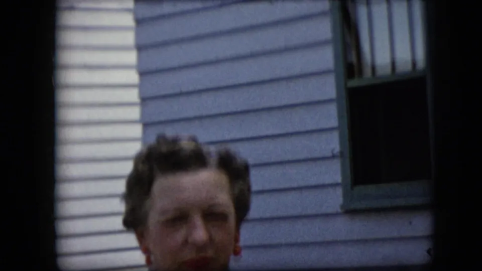 The woman is framed lower in the 1969 California home-movie shot, with her face near the bottom edge and a green-trimmed window above on the sided wall.