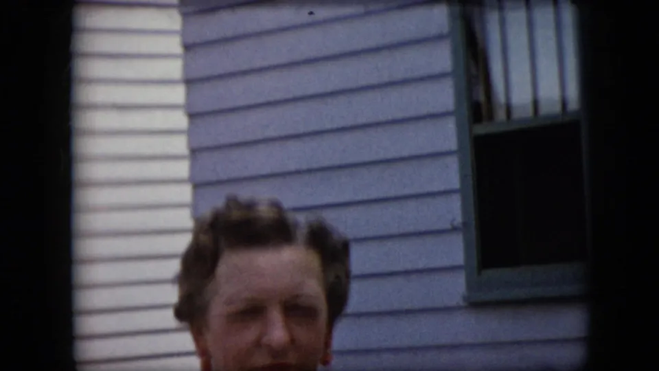 A mature woman remains partly cropped near the lower edge of this 1969 California 8mm frame, with light horizontal siding, green window trim, and dark transfer borders visible.