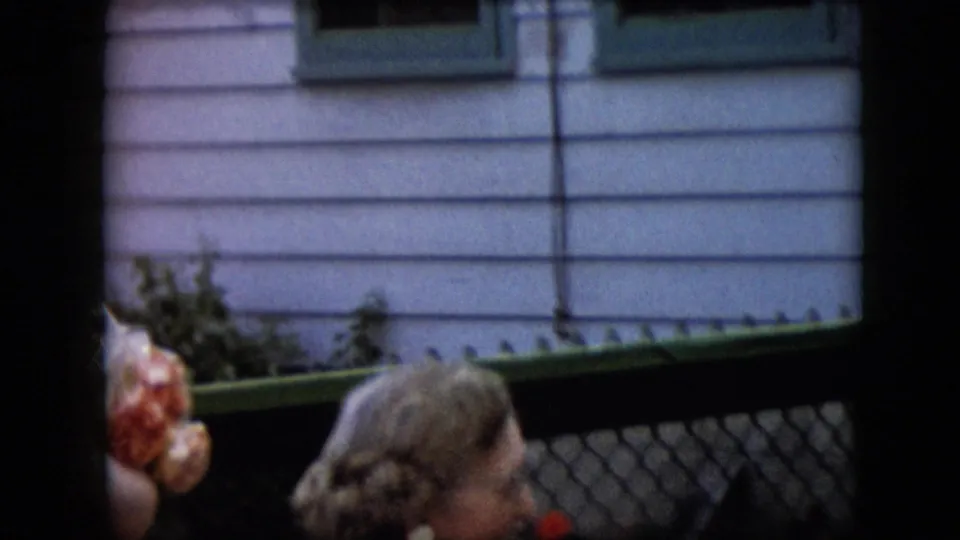 A partial outdoor home-movie frame shows the lower edge of the woman or another adult near a chain-link fence, greenery, pale siding, and a dark screened window.