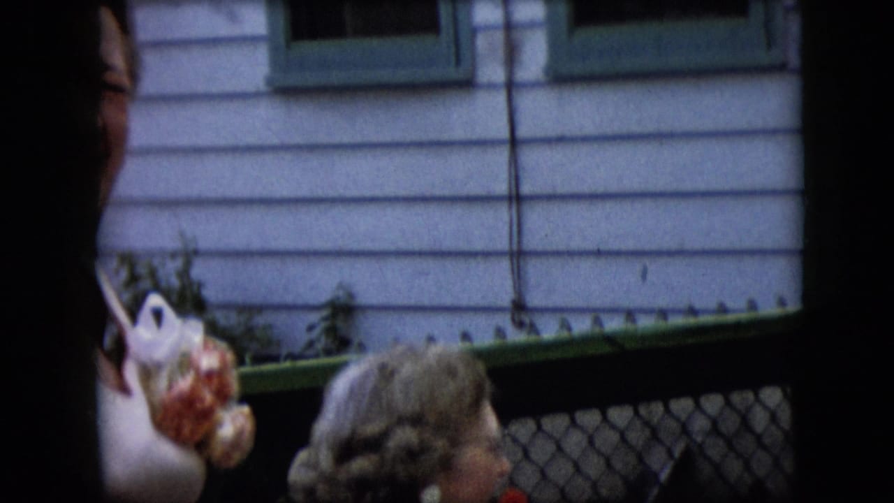 Two women in formal attire near a chain-link fence and clapboard house, partly obscured by dark foreground shapes.
