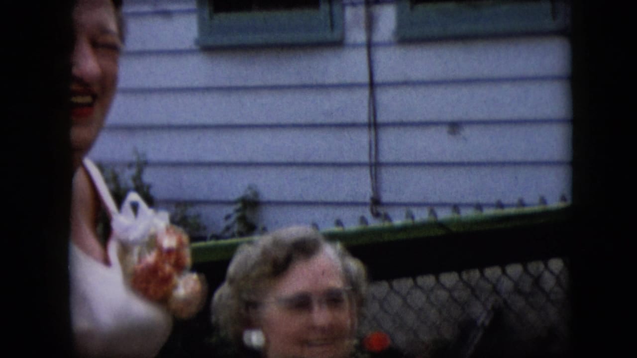 Smiling woman in a white dress holding a wrapped item beside an older woman near a chain-link fence.