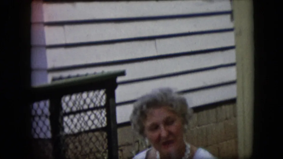 1969 California frame showing an older woman with styled hair and light clothing near suburban clapboard siding and a chain-link fence during the gathering.