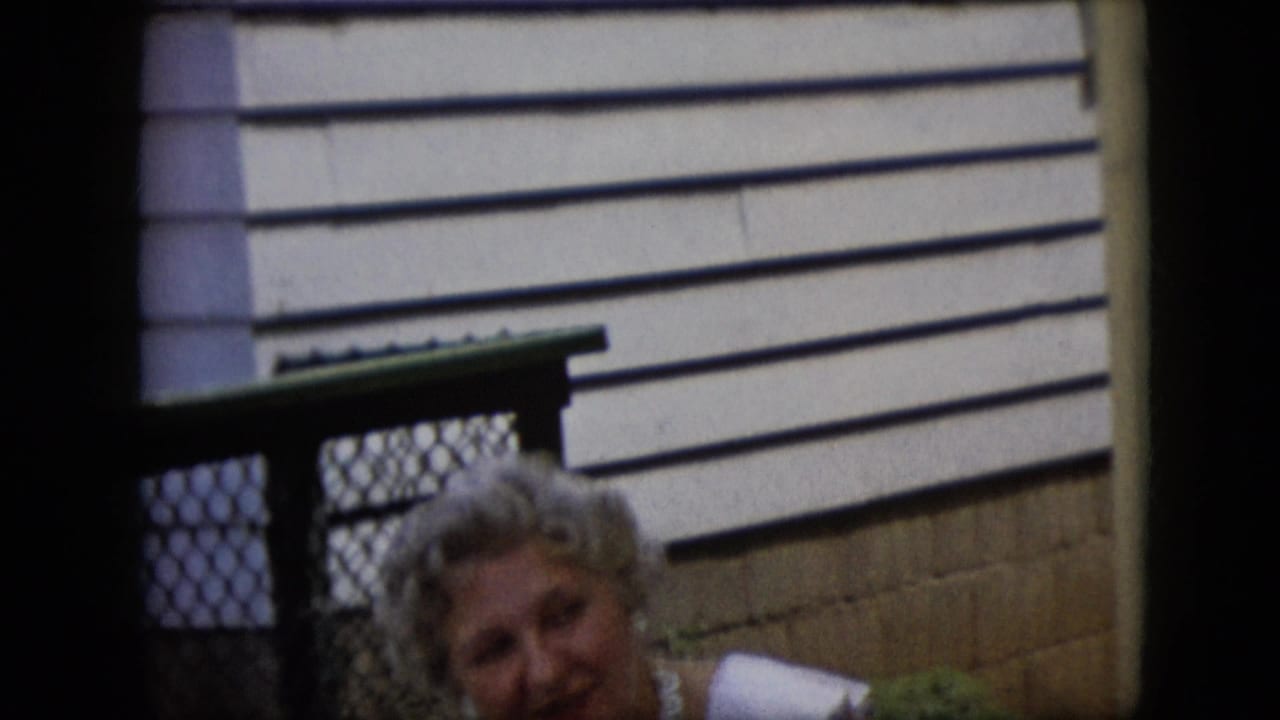 In 1969 California home-movie footage, an adult woman in a white sleeveless dress and pearl necklace is partly visible near a chain-link gate and sunlit horizontal siding.