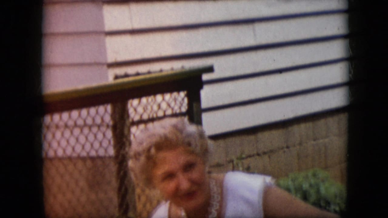Woman in white dress looks toward the camera area near a chain-link gate and house exterior.
