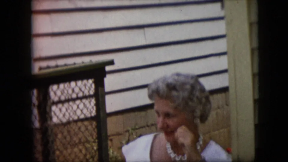 In the 1969 home-movie frame, the woman in pearls rests her chin on her hand beside the chain-link gate and house siding, suggesting a candid conversational pause.