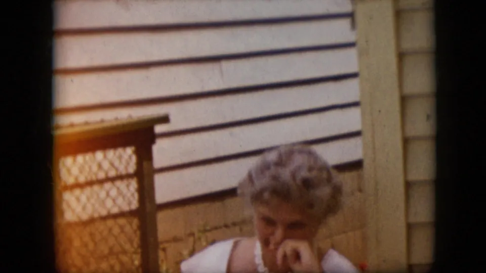 The adult woman in a white dress and pearl necklace looks downward near the chain-link gate, with warm 8mm exposure and the house exterior behind her.