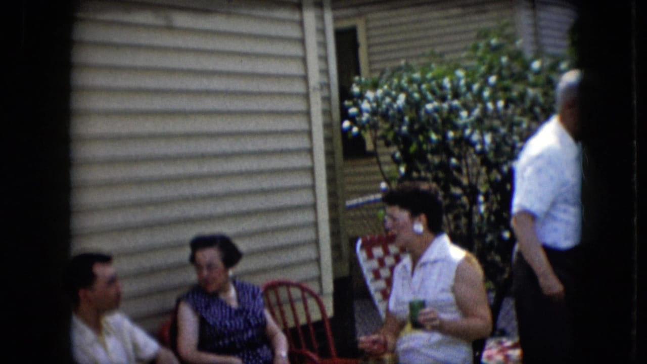 California 1969 home-movie frame of adults relaxing outside a beige-sided house, with red patio chairs, a checkered chair, drinks, and a flowering bush visible.