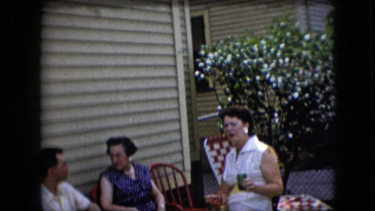 Three adults sit on patio chairs beside a flowering bush and beige house siding.