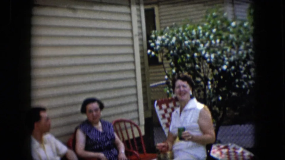 California 1969 home-movie frame of three adults seated outdoors, with one woman smiling toward the camera while holding a cup near the side of a house.